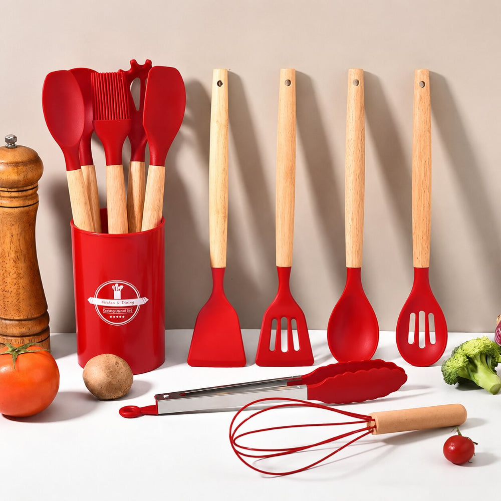 A 7-piece silicone kitchen utensil set with red heads and light wood handles, displayed on a white surface with various food props and a wooden pepper mill against a beige background.