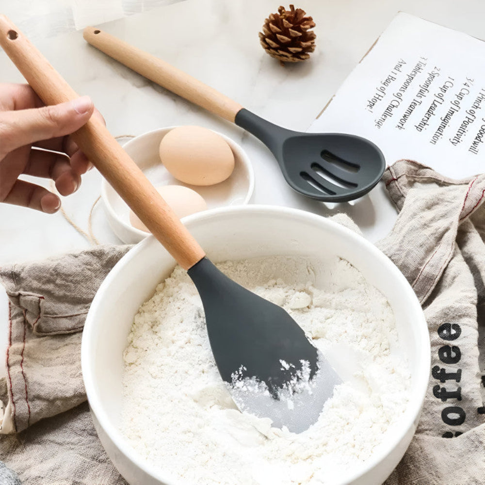 Person using a black silicone spatula to mix flour in a bowl with a wooden handle.