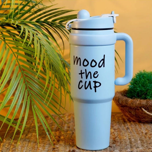 A light blue insulated travel mug with a handle and lid, featuring the phrase 'mood the cup' printed in black script. The mug is styled on a woven mat with a yellow background, green palm leaves, and a small potted plant.